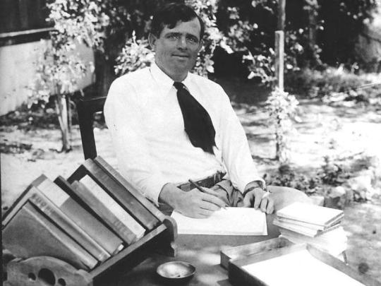 Man wearing a white shirt and black tie, sitting outside..  He is writing, there are books on the table.