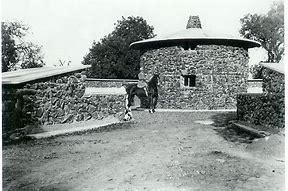 Woman on a hourse stanging in front of a round stone building.